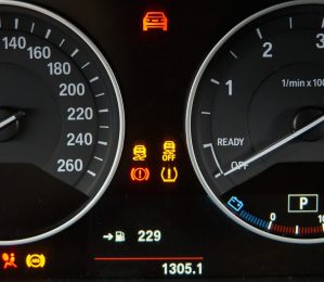 A wide shot of a modern car's dashboard, featuring a digital display in the center showing an open car door icon. Analog gauges for RPM and speed are visible, along with several dashboard warning lights illuminated, such as the engine light and a low fuel indicator.