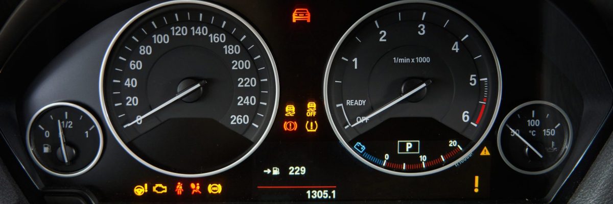 A wide shot of a modern car's dashboard, featuring a digital display in the center showing an open car door icon. Analog gauges for RPM and speed are visible, along with several dashboard warning lights illuminated, such as the engine light and a low fuel indicator.