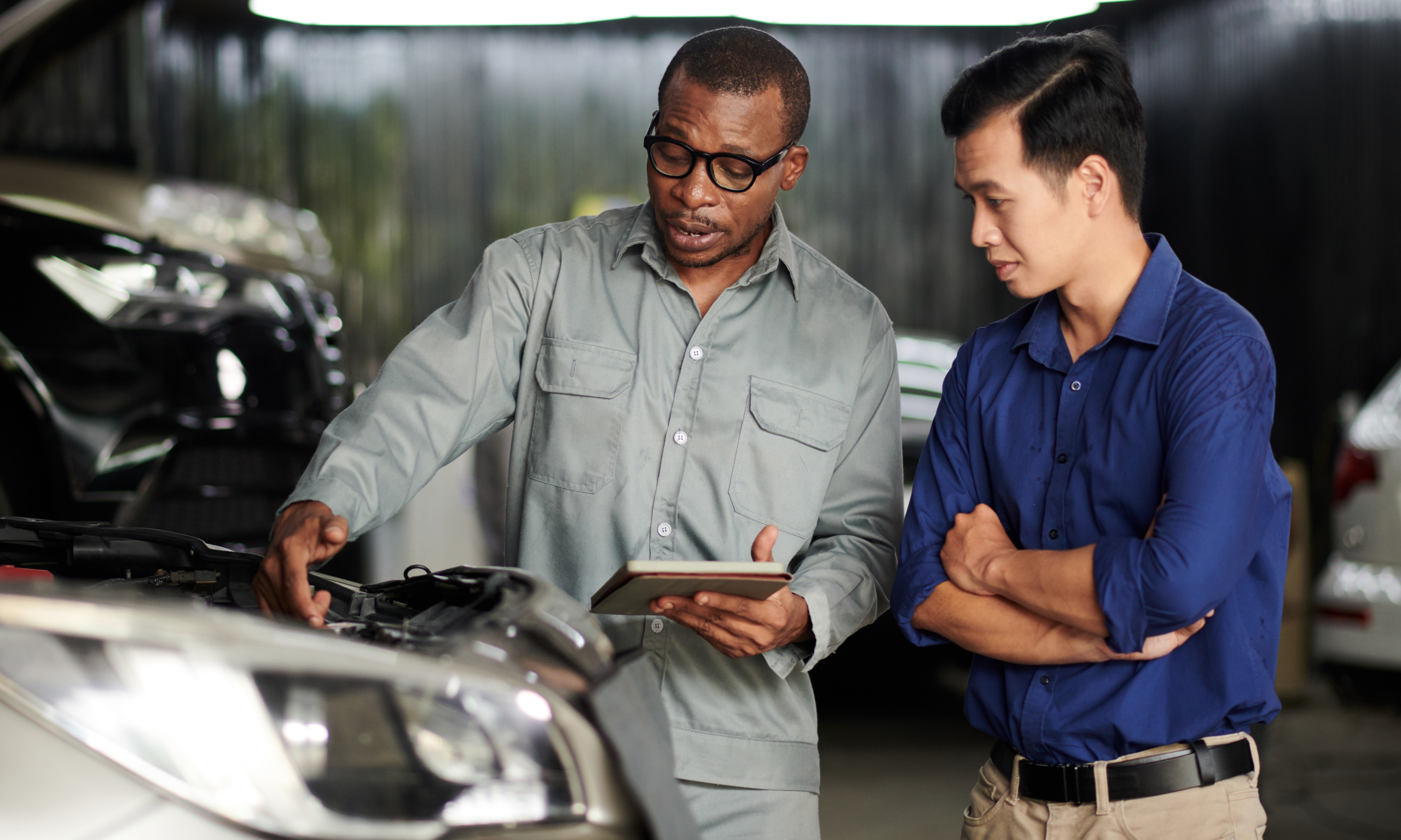 A mechanic at service station explains issues to a customer using a tablet while standing beside a car with the hood open.