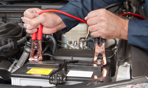 A person performs a battery check by connecting red and black jumper cables to the battery terminals to jump-start the vehicle.