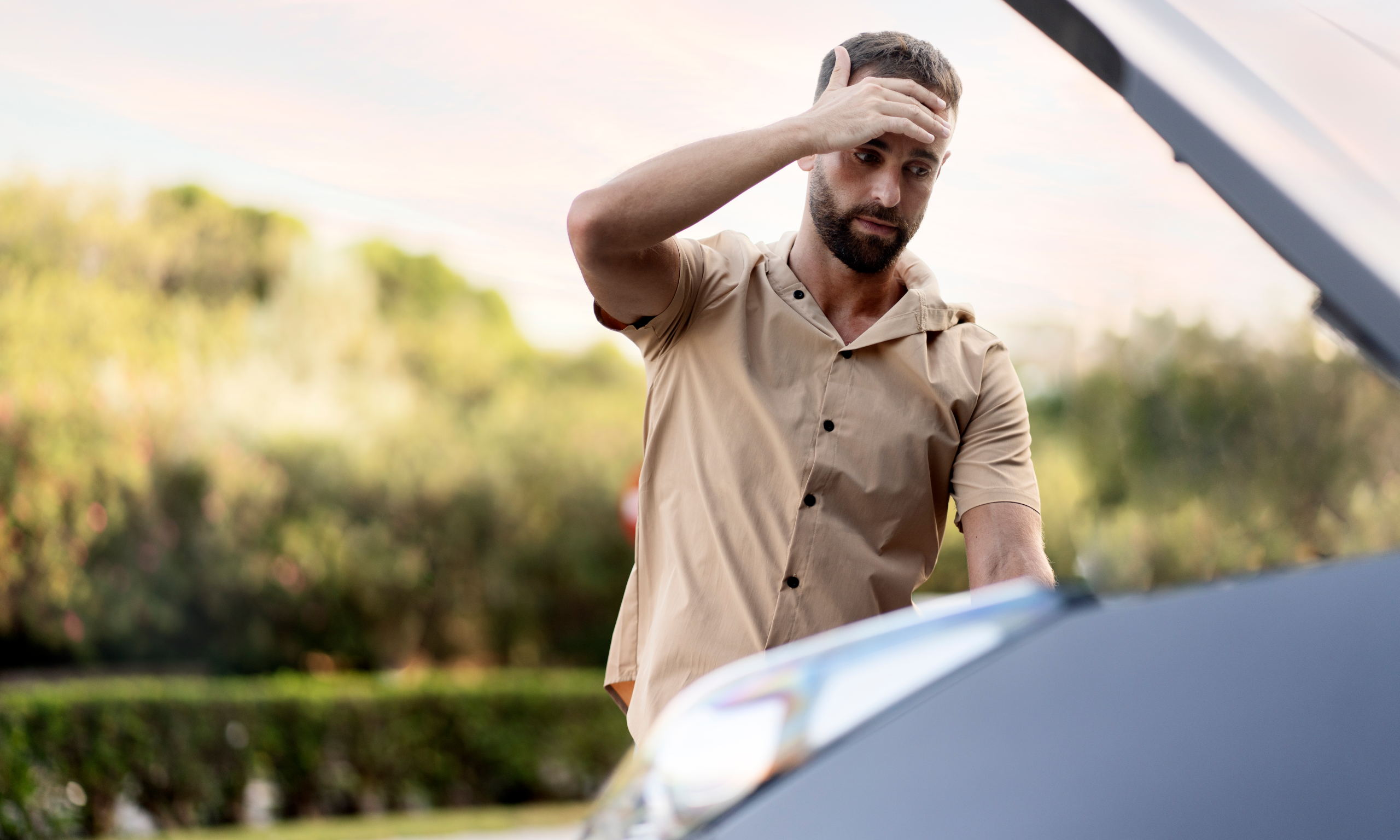 A concerned man stands in front of a broken-down car with the hood open.
