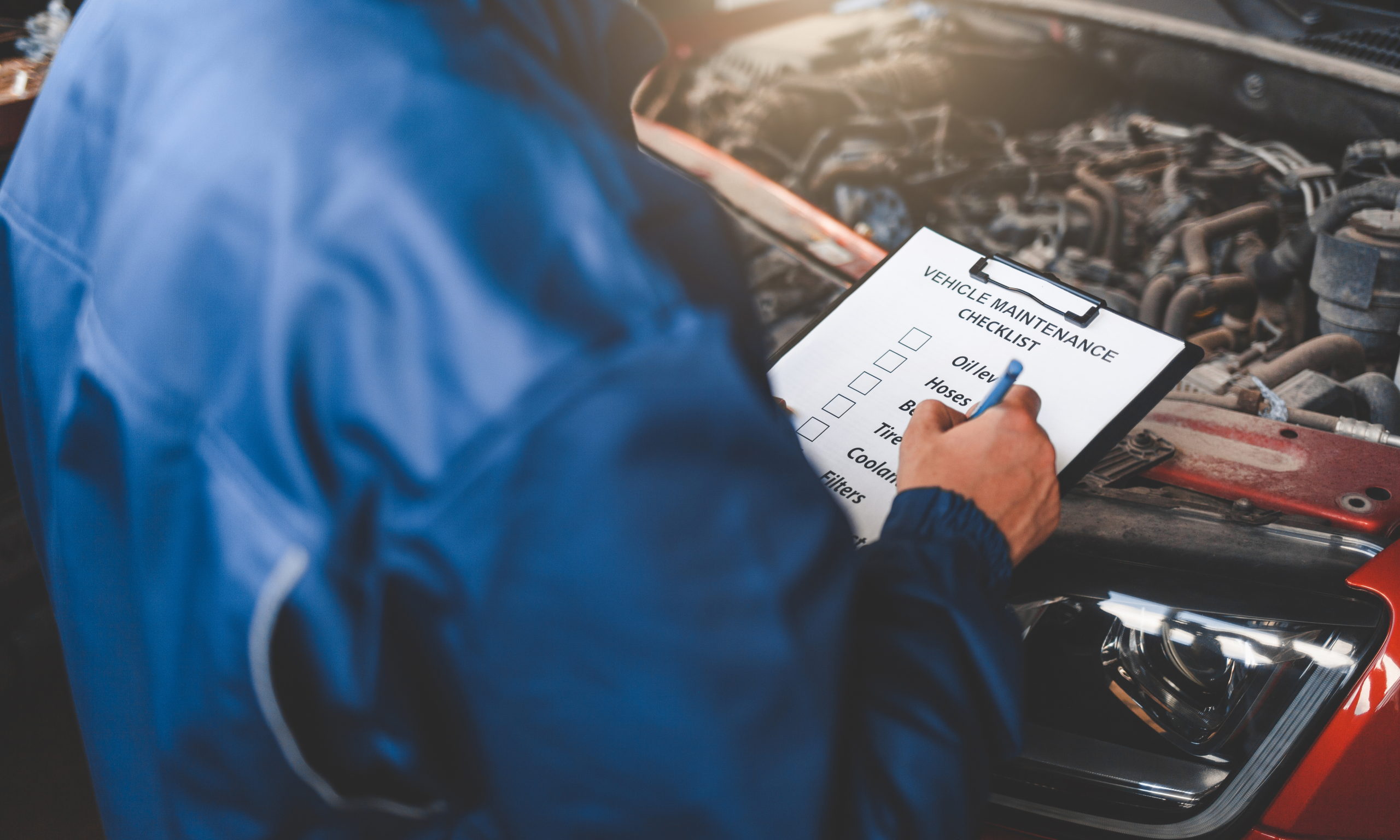 A mechanic in a blue uniform holding a checklist labeled “Vehicle Maintenance” while inspecting an open car hood at Acton Service Centre, possibly reviewing cambelt condition.