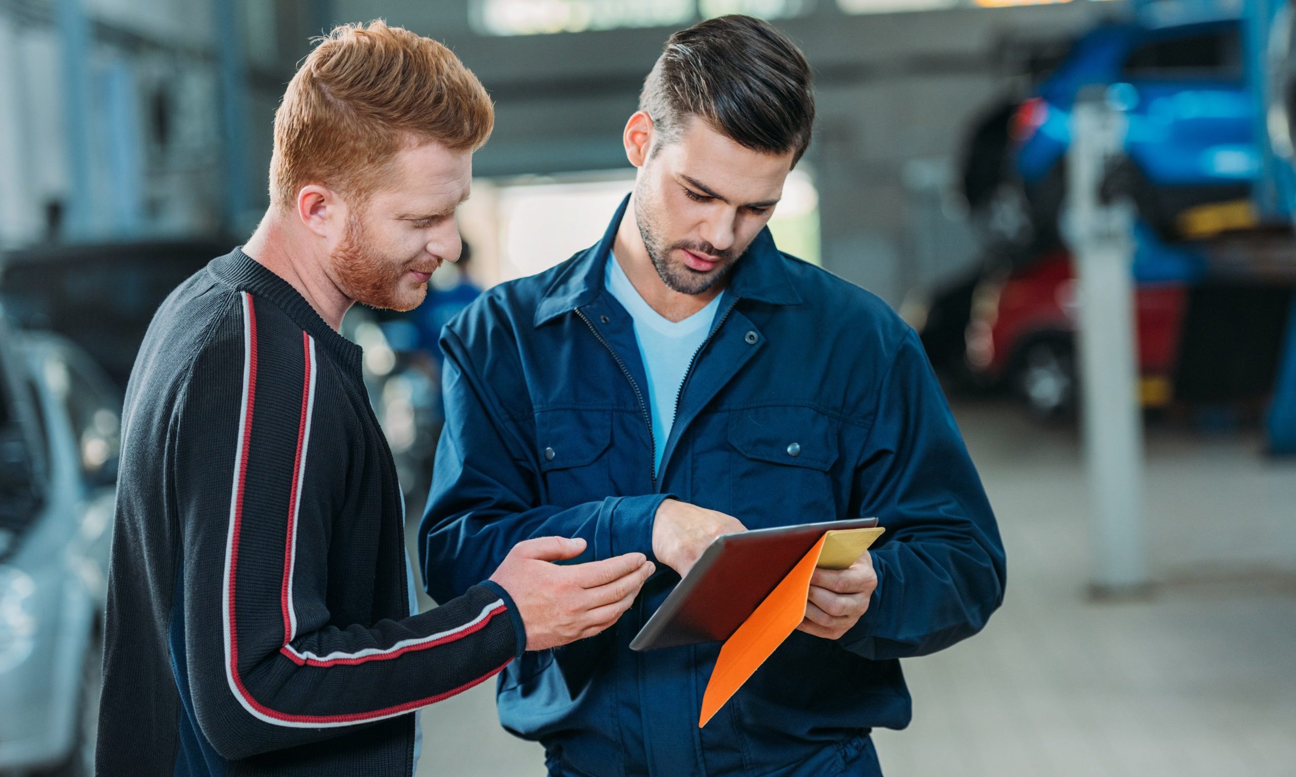 A mechanic shows a customer repair cost details on a tablet. They are standing in a busy auto workshop with vehicles in the background.
