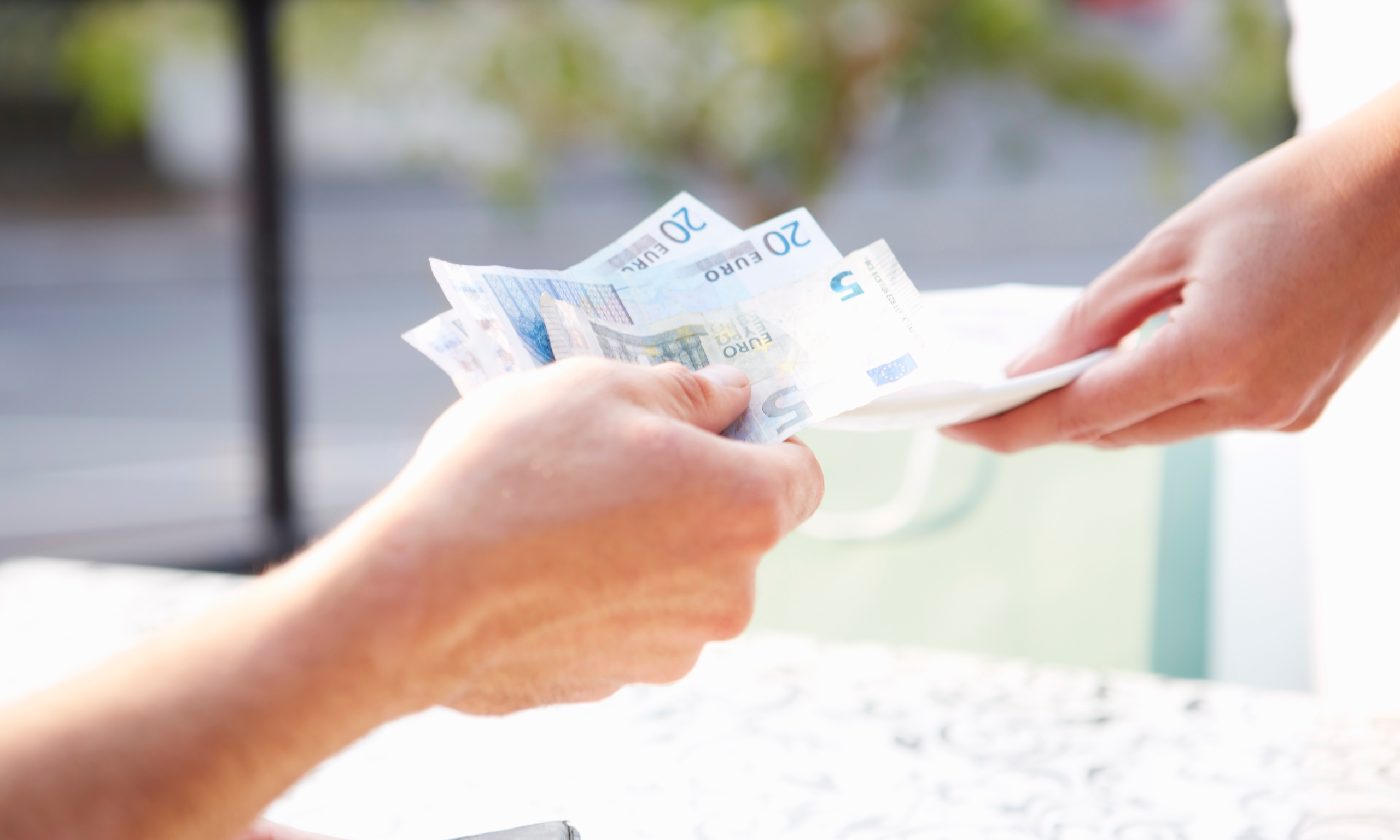 A customer hands over euro bills to make a payment after a service. The transaction takes place at a service desk with a soft background blur.