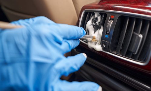 A person wearing blue gloves using a brush to clean a car's air con vent with foam, showing a method to remove air con smell.