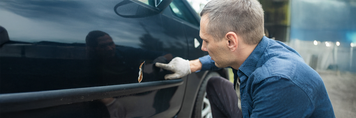 A man in a blue shirt and white gloves inspects a significant rust-colored scratch on a black car door, highlighting a car scratch repair problem.