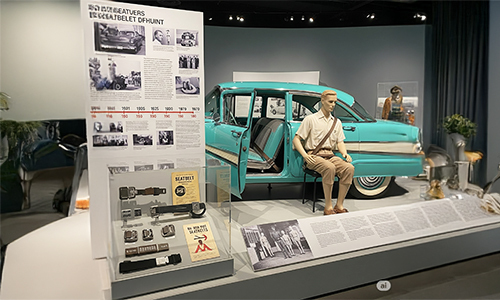 An exhibit on the history of car safety, featuring a vintage blue car with a mannequin wearing a seatbelt. A display board shows old photos and different types of seatbelt buckles from over the years.