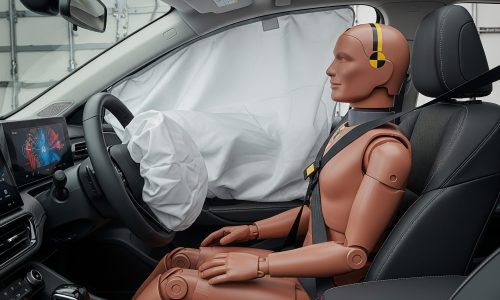 A crash test dummy sits in the driver's seat of a modern car. An airbag has deployed from the steering wheel.