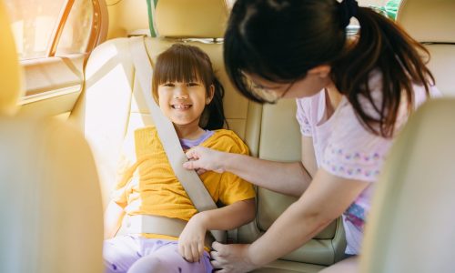 A mother is helping a young girl in the back seat of a car fasten her seatbelt, showing how to buckle up.