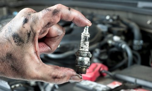 A mechanic’s hand holding a dirty, used spark plug with visible grease stains, with a car engine blurred in the background.