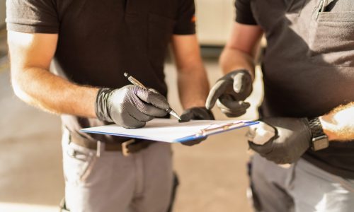 Two auto service technicians wearing gloves and uniforms discussing and writing notes on a clipboard in a brightly lit garage.