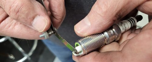 A mechanic is holding a spark plug in focus, with a vehicle’s hood open and the engine out of focus in the background.