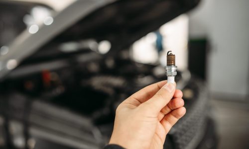 A mechanic is holding a spark plug in focus, with a vehicle’s hood open and the engine out of focus in the background.